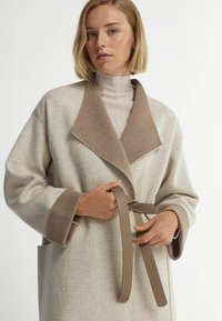 Ljusbeige kappa med sjalkrage och bruna detaljer. Har stora fickor, lös passform och ett knytbälte. Tillverkad av mjukt, strukturerat tyg.