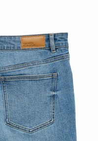 Pantalones vaqueros de mezclilla azul claro con un parche de cuero marrón que lleva la inscripción "VERO MODA WE DO DENIM." La tela tiene una textura sutil y detalles de costuras visibles.