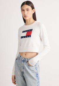 Krátky biely sveter s rebrovanými manžetami, s centrálnou modrou a červenou nášivkou s nápisom "TOMMY JEANS." Nosený s svetlomodrými džínsami.