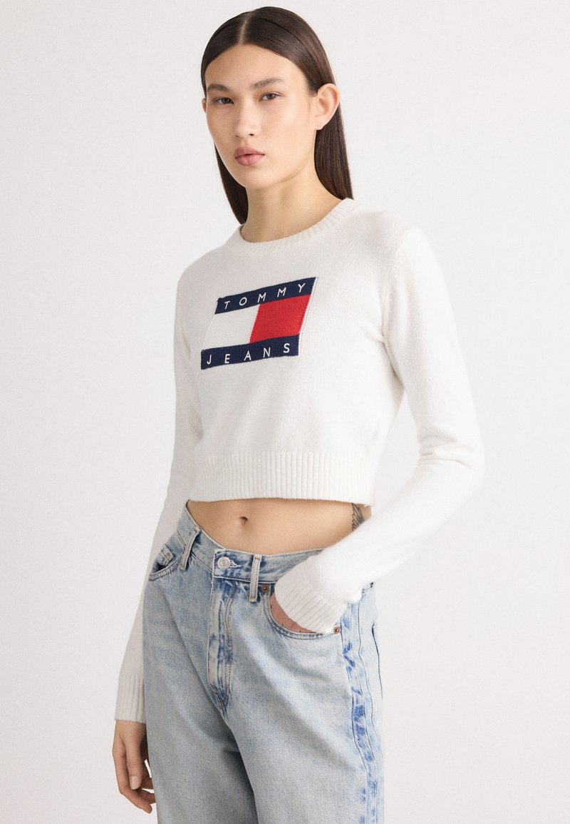Krátky biely sveter s rebrovanými manžetami, s centrálnou modrou a červenou nášivkou s nápisom "TOMMY JEANS." Nosený s svetlomodrými džínsami.