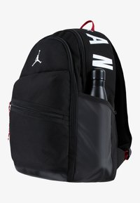 Rucsac negru cu material neted, dotat cu un buzunar frontal cu fermoar, suport vizibil pentru sticla de apă și accente roșii cu branding alb.