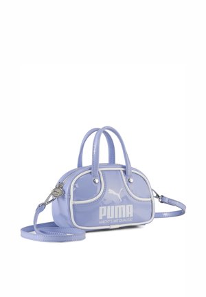 Mujer con chaqueta Puma en blanco y negro, gafas de sol negras, pendientes de aro plateados, pulseras y sosteniendo un pequeño bolso Puma de color lila claro.