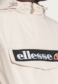 Ljusbeige vindjacka med hög krage, zippered stängning och en framträdande svart patch med "ellesse" logotyp. Texturerad tyg.