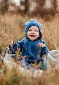 Sininen fleecetakki, jossa on paneelisuunnittelu ja korva-tehosteet, istuu vilttiin kääritty vauva ruohikolla, hymyillen ja pitäen ruhoa kädessään.