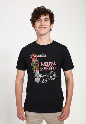 Jonge man lacht, draagt een zwart T-shirt met een rood, wit en groen embleem en tekst van het Mexicaanse voetbalteam uit 1986.