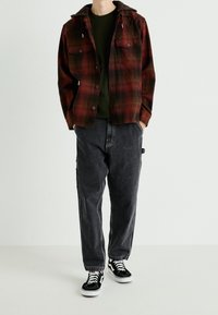 Camicia di flanella a scacchi rossa e nera con cappuccio, abbinata a pantaloni larghi grigi scuri e sneakers nere con suola bianca. Stile casual.