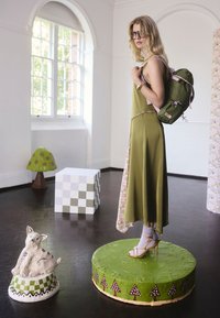 Olivovo zelené šaty s kvetinovými detailmi, skombinované s ružovo-zeleným batohom. Predstavené na zelenom podstavci, vedľa ktorého sa nachádza dekoratívne keramické zviera.