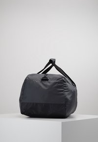 Musta nailoninen urheilukassi, jossa on kaksinkertaiset kahvat ja klipsiyksityiskohta, asetettuna valkoiselle jalustalle yksinkertaista harmaata taustaa vasten.