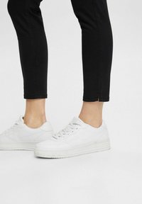 Zapatillas blancas con un acabado texturizado, que presentan un diseño minimalista, forma de bajo y una suela gruesa y estriada. Llevadas con pantalones negros recortados.