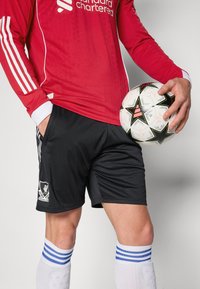 Maillot de football rouge à manches longues avec des accents et un logo blancs, associé à un short noir portant un logo. Chaussettes rayées blanches et bleues. Tenant un ballon de football à motifs.
