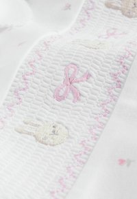 Hvidt stof med tekstureret vævning har lyserød broderi af en sløjfe, en kanin og små blomsterdetaljer. Blødt og delikat udseende.