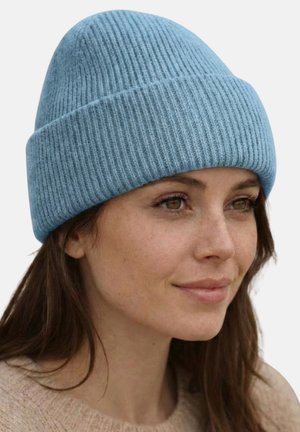 Junge Frau mit hellblauer gestrickter Mütze und beigem Pullover, die leicht nach rechts schaut, mit einem sanften Lächeln und natürlichem Make-up.