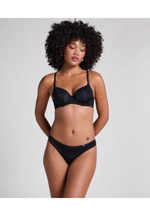 Vrouw met krullend haar poseert in een zwarte beha en bijpassende zwarte slip tegen een egale lichte achtergrond.