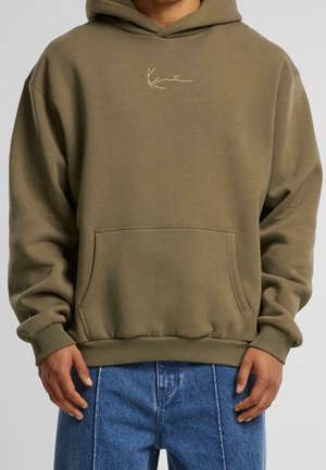 Persoon draagt een olijfgroene hoodie met een voorvak en een klein beige logo, gecombineerd met een blauwe spijkerbroek, staand tegen een neutrale achtergrond.