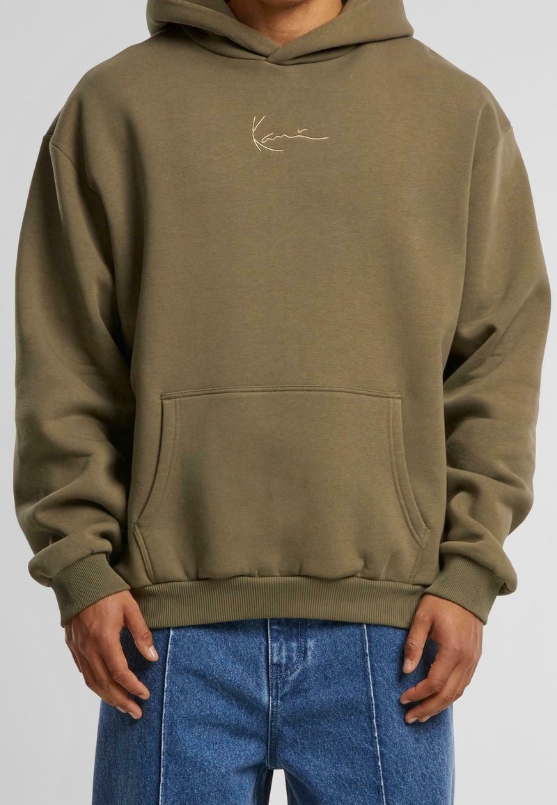 Personne portant un sweat à capuche vert olive avec poche avant et petit logo beige, associée à un jean bleu, debout devant un fond neutre.