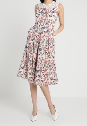 Hihaton valkoinen midimekko monivärisellä kukkakuviolla, pitsiyksityiskohta pääntiellä, yhdistetty valkoisiin kärjestä teräviin korkokenkiin.