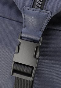 Detail batohu z námořnické látky s černou plastovou sponou a nastavitelným popruhem, s texturovaným povrchem a kontrastním prošitím.