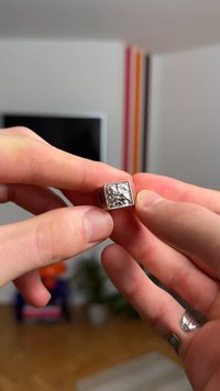 Anillo de plata con una cara cuadrada texturizada que muestra un patrón detallado. El anillo tiene un acabado liso y pulido y se sostiene entre los dedos.