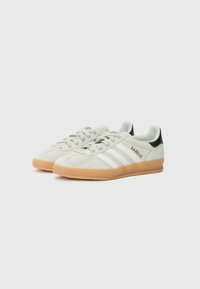 Adidas Gazelle sneakers with a light grey suede upper, white stripes, gum rubber sole, and black heel accent. Durable and classic design.