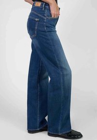 Jeans en denim bleu à taille haute et jambes larges, dotés de cinq poches et d'une patch en cuir marron à l'arrière. Des détails cousus mettent en valeur la texture du tissu.