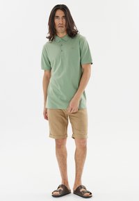 Grön bomullspiké med krage, parat med tanfärgade uppvikta shorts och bruna sandaler. Enkelt mönster med en subtil logotyp på bröstet.