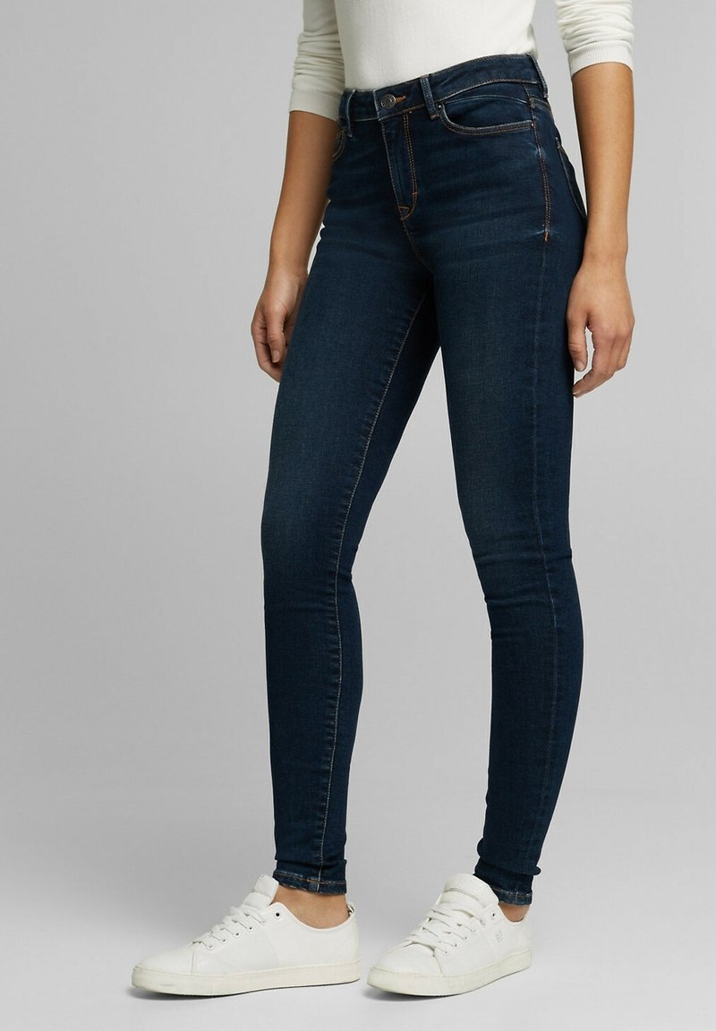 Personne portant un jean skinny bleu foncé, un haut blanc à manches longues et des baskets blanches, debout devant un fond gris uni.