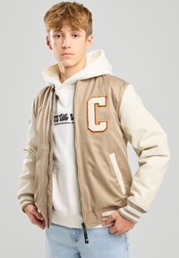 Beige bomber jacket with white leather sleeves, featuring a felt letter "C" patch. Ribbed cuffs and a zip closure. Layered over a hoodie.