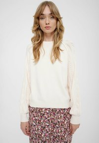 Maglione lavorato a maglia crema con dettagli in pizzo sulle maniche, polsini e orlo a coste. Abbinato a una gonna con stampa floreale che presenta tonalità di viola e rosa.