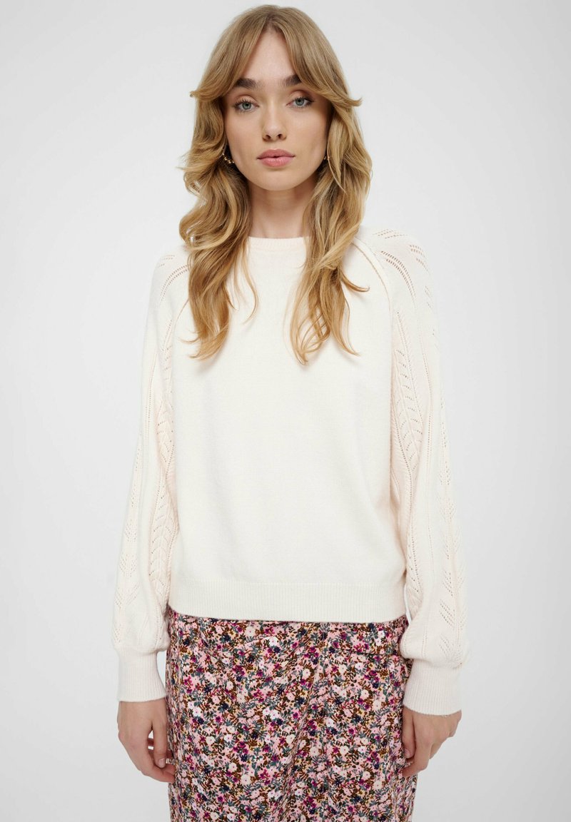 Maglione lavorato a maglia crema con dettagli in pizzo sulle maniche, polsini e orlo a coste. Abbinato a una gonna con stampa floreale che presenta tonalità di viola e rosa.