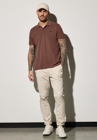 Camisa polo de algodón marrón con cuello, chinos beige y zapatillas blancas. Mangas cortas y un logo sutil en el lado izquierdo. La gorra es color crema.