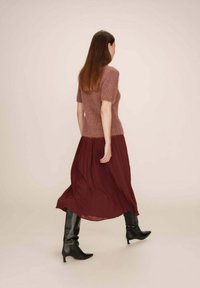 Un haut tricoté rose à manches courtes avec une texture douce, associé à une jupe plissée bordeaux fluide et des bottes en cuir noir jusqu'aux genoux.