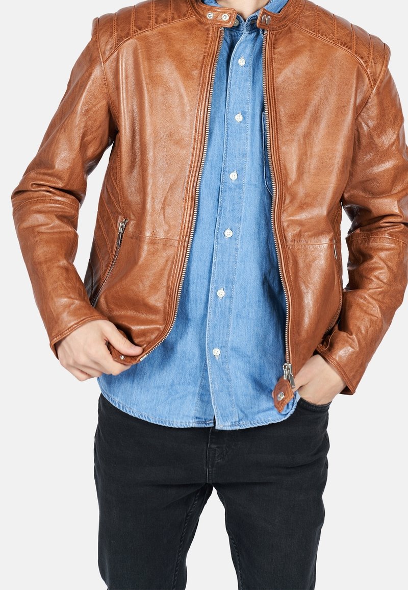 Braune Lederjacke mit einem Reißverschluss, gesteppten Schulterakzenten und Seitentaschen, getragen über einem Jeanshemd, kombiniert mit schwarzen Jeans.