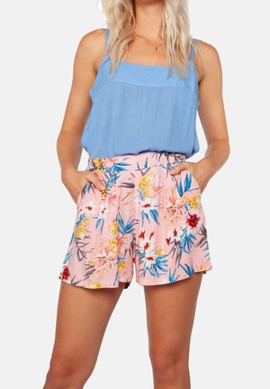 Femme portant un haut bleu sans manches et un short rose à fleurs, les mains dans les poches, sur un fond uni.