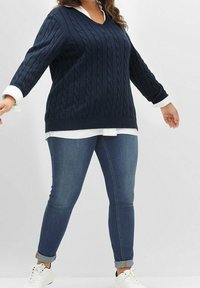 Osoba nosząca granatowy sweter w warkocze na białej koszuli, niebieskie dżinsy z podwiniętymi nogawkami i białe trampki, stoi z lekko wyciągniętymi ramionami.
