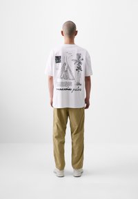 White graphic t-shirt featuring black prints of trees and designs on the back, paired with beige trousers and light-colored shoes.