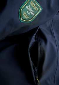 Melnas āra jakas ar zaļu un zeltītu izšūtu logotipa uzlīmi un kabatu ar melnu rāvējslēdzēju. Gluda auduma tekstūra.