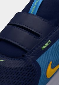 Κοντινό πλάνο ενός μπλε αθλητικού παπουτσιού με κίτρινο λογότυπο Nike, λουράκια βέλκρο και το κείμενο "Dynamic Fit" στη μεριά.