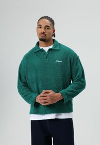 Zachte groene trui met een kraag, twee knopen en geborduurd logo. Lange mouwen en een ontspannen pasvorm, gedragen over een wit overhemd.