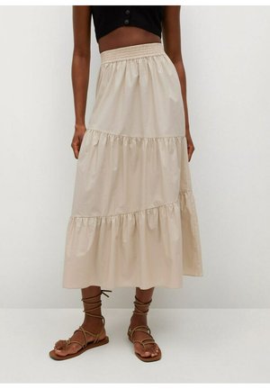 Femme portant une jupe midi beige à volants, un top court noir et des sandales plates marron à brides, debout devant un fond uni.