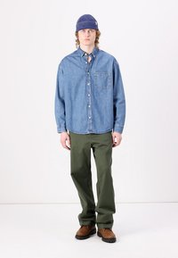 Cămașă din denim cu butoni, de culoare bleu deschis, pantaloni verzi cu talie înaltă, cizme maro și o căciulă albastră din tricot, toate pe un fundal neutru.