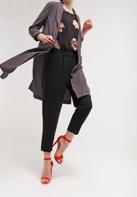 Cardigan largo gris, blusa negra con estampado floral, pantalones negros y sandalias de tacón alto rojas. Patrón floral notable en la blusa; silueta moderna y elegante.