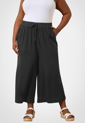 Woman wearing black wide-leg cropped pants with drawstring waist, white tank top, and white wedge sandals on plain background.