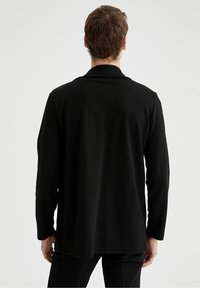 Svart, långärmad jacka med fiskbensstruktur. Har en notch-lapel och en lös passform som slutar vid låren. Minimalistisk design.