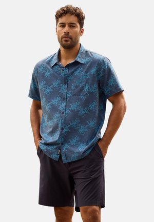 Hombre con una camisa de botones de manga corta azul con estampado floral y pantalones cortos negros, de pie con las manos en los bolsillos frente a un fondo liso.