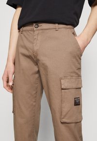 Hombre que lleva pantalones cargo beige con bolsillos con solapa y una camiseta negra de mangas cortas, con una mano en el bolsillo, cortados a la altura de la cintura y los muslos.