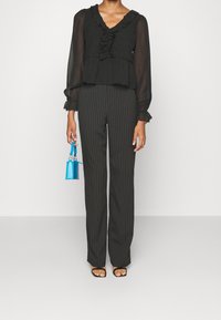 Blusa nera con volant e maniche lunghe, abbinata a pantaloni neri a righe sottili. Gli accessori includono una piccola borsa blu e sandali neri.