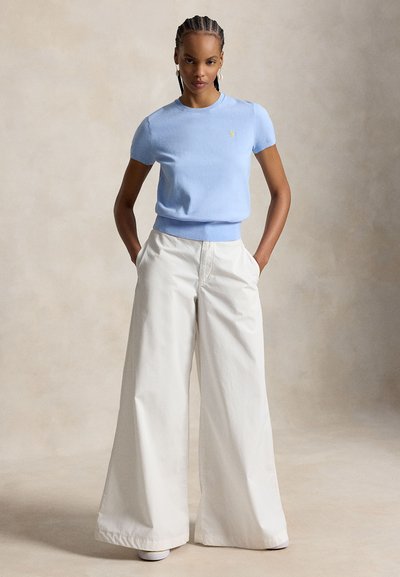 Γυναίκα με πλεξουδάκια, που φοράει ανοιχτό μπλε πλεκτό τοπ με κοντά μανίκια και λευκό παντελόνι ψηλόμεσο, φαρδύ στο κάτω μέρος, με τα χέρια στις τσέπες.