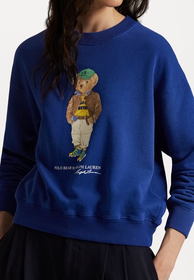 Blauwe sweatshirt van zacht materiaal, met een beerafbeelding in een bruine jas en groene pet, voorzien van een geribbelde halslijn en manchetten.