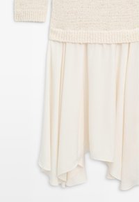 Crèmekleurige gebreide trui met geribde zoom, gecombineerd met een vloeiende, asymmetrische crèmekleurige rok.