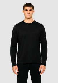 Maglietta nera a maniche lunghe realizzata in tessuto morbido, con scollatura rotonda e un branding discreto sul petto, indossata con pantaloni neri.